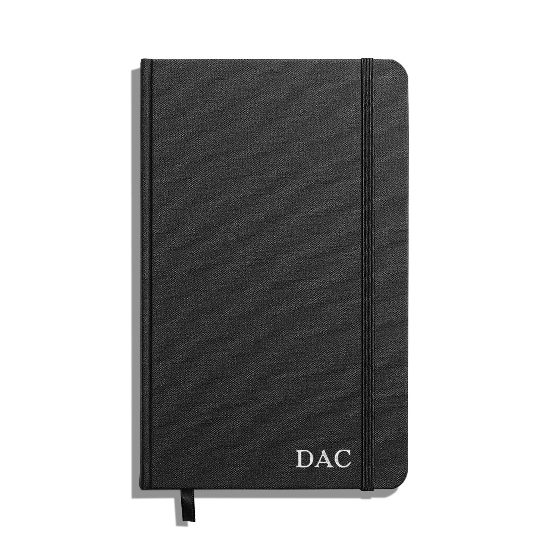The Shinola Medium Hard Ruled Journal in black features a hardcover, elastic band closure, ribbon bookmark, and acid-free paper for lasting quality, with "DAC" initials printed in white on the bottom right corner.