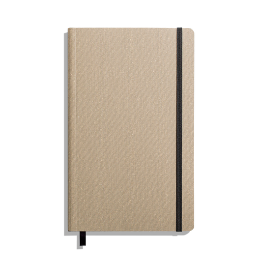 The Shinola Medium Soft Ruled Journal - Oat is shown closed with a textured beige cover, black elastic band on the right, and a black ribbon bookmark against a white background. Features durable smyth-sewn binding.