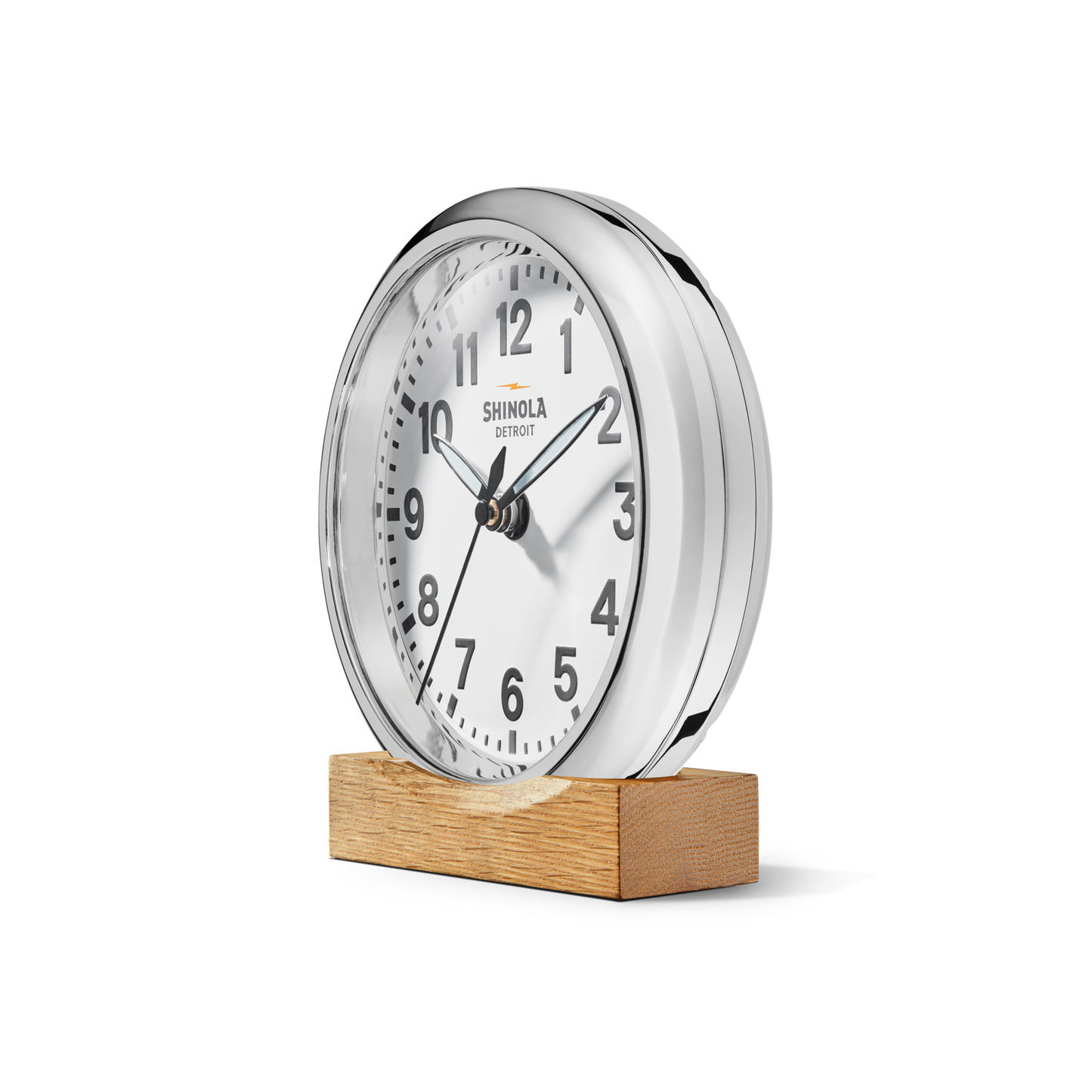 A round, silver 6" Runwell Clock - White by Shinola features a white face with black numbers and hands, sits on a rectangular wooden base, and displays the time as 2:12.
