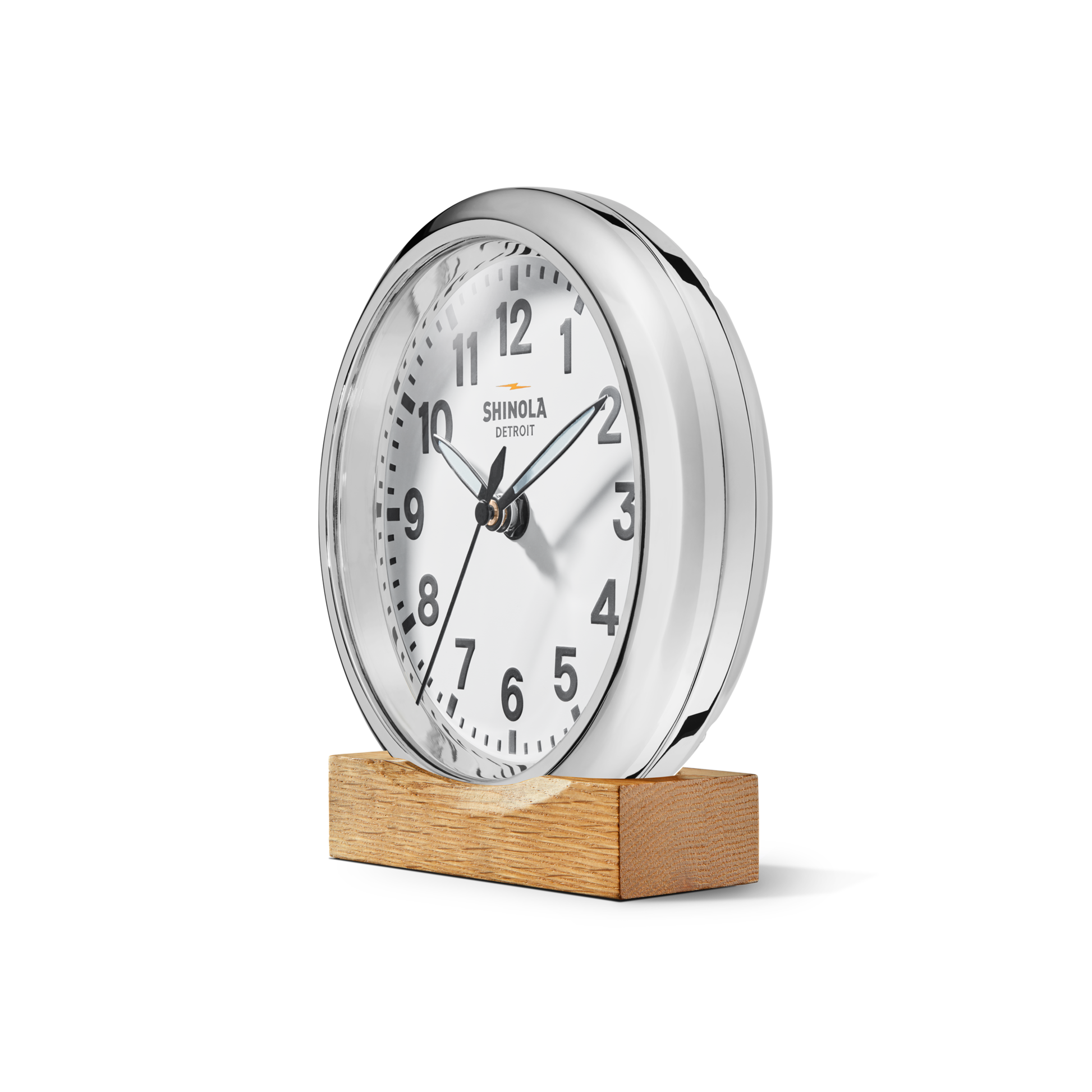 A round, silver 6" Runwell Clock - White by Shinola features a white face with black numbers and hands, sits on a rectangular wooden base, and displays the time as 2:12.