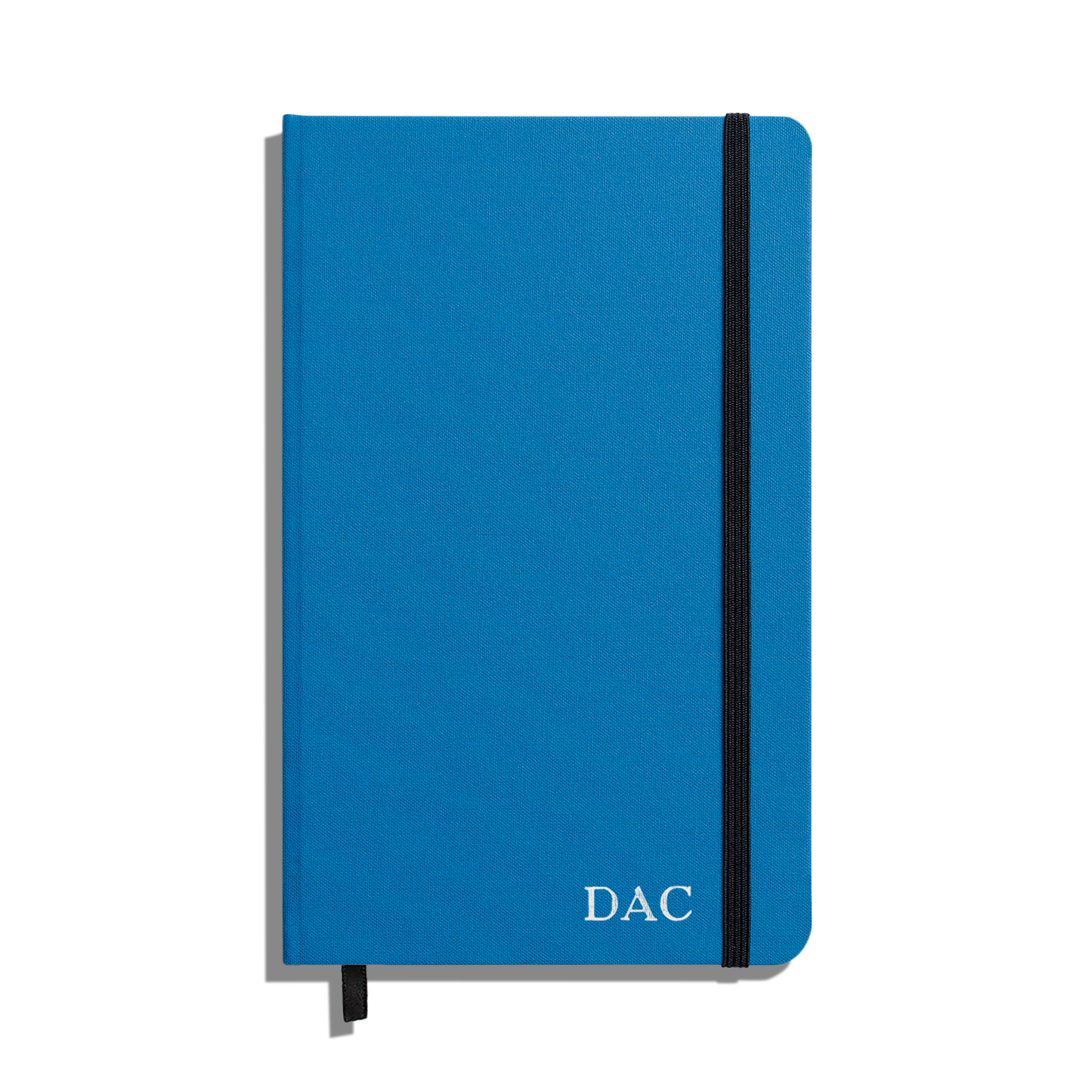 The Shinola Medium Hard Ruled Journal in cobalt features a closed blue linen cover, "DAC" in white on the bottom right, smyth-sewn binding, a black elastic band closure, and a black ribbon bookmark peeking from the bottom.