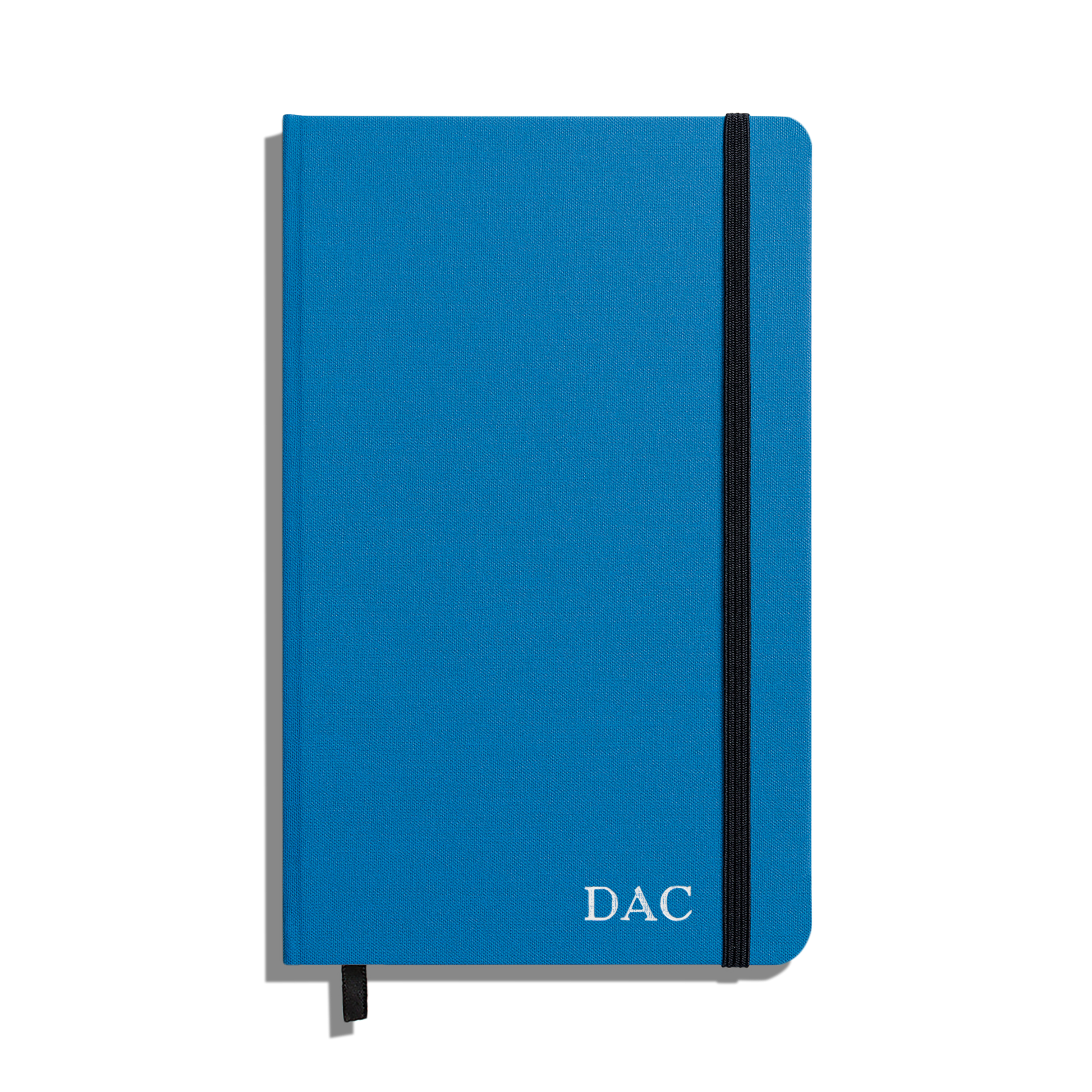 The Shinola Medium Hard Ruled Journal in cobalt features a closed blue linen cover, "DAC" in white on the bottom right, smyth-sewn binding, a black elastic band closure, and a black ribbon bookmark peeking from the bottom.