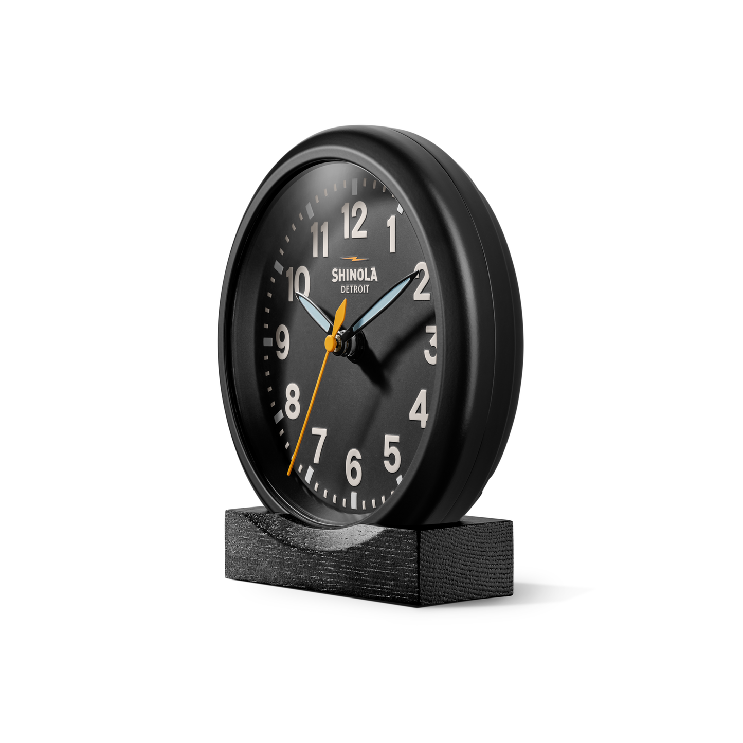 The 6" Runwell Clock - Black by Shinola features a round black analog face with white and orange hands, a yellow second hand, and white numbers. It sits on a black rectangular base against a plain white background. Inspired by Detroit watchmaking.