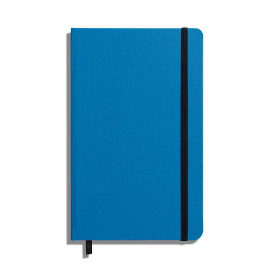 The Shinola Medium Hard Ruled Journal - Cobalt is a closed blue hardcover notebook with acid-free paper, a vertical black elastic band, and a matching ribbon bookmark, shown on a white background.