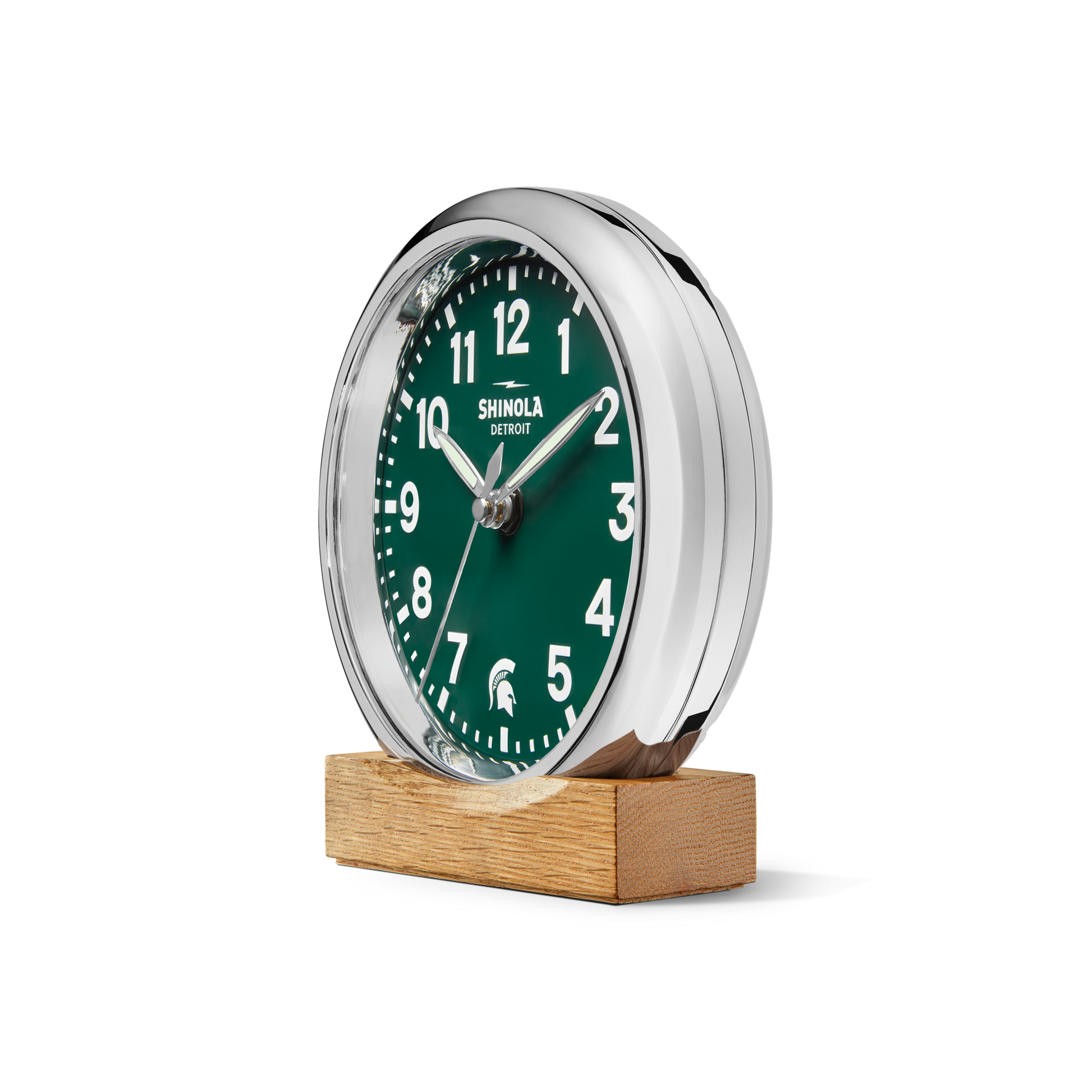 A 6" Runwell Clock - Green by Shinola, featuring a round green face with white numbers and hands in a silver frame, is displayed on a rectangular wooden base against a plain white background.
