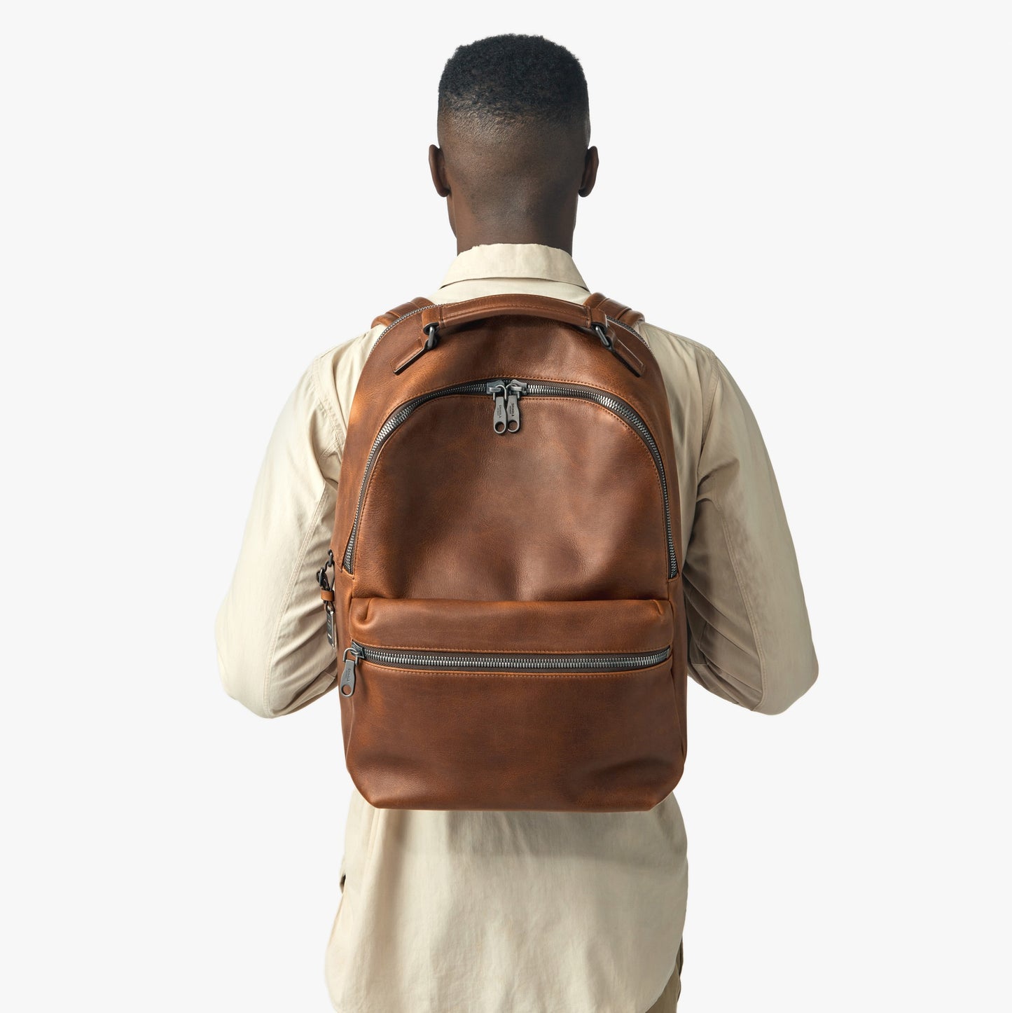 A person in a beige shirt faces away, displaying the Shinola Runwell Backpack in Medium Brown with multiple zippered compartments, set against a plain white background.
