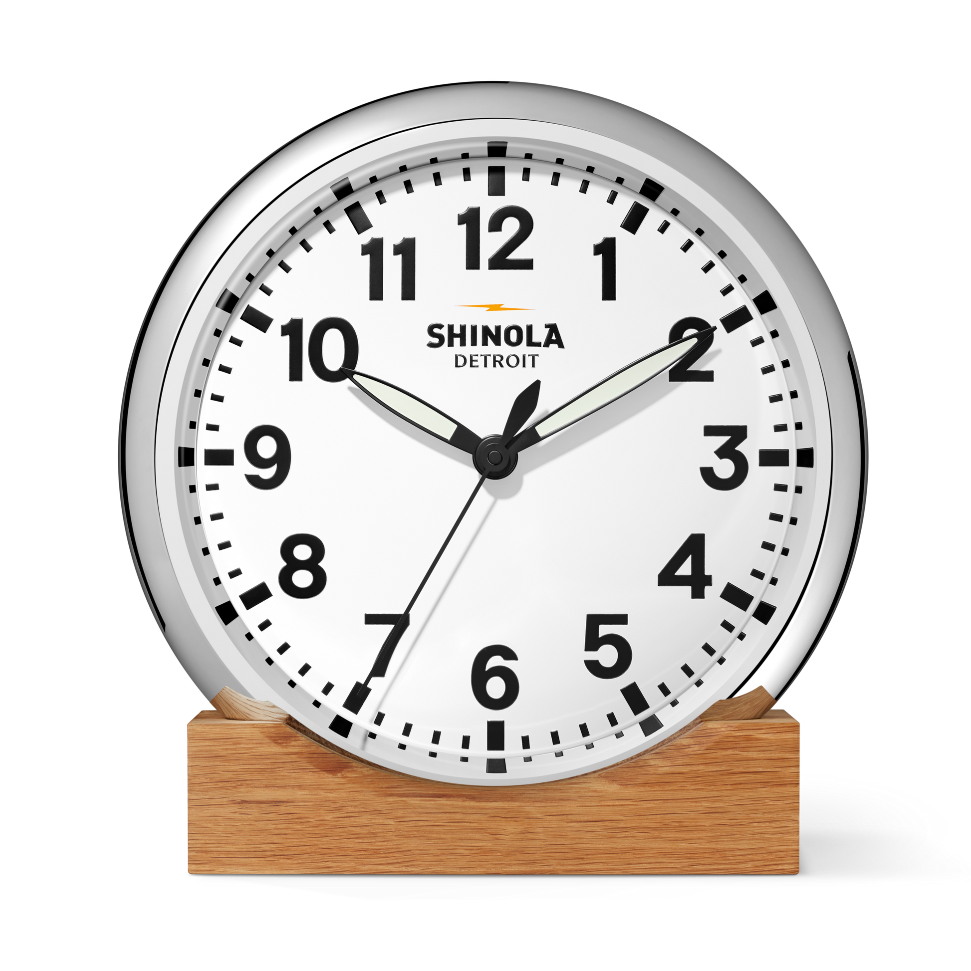 A 14" Shinola Runwell Clock with a white face, black numbers, and a silver frame sits on a wooden base. The clock shows 1:51:34, with the Shinola brand printed on the face.