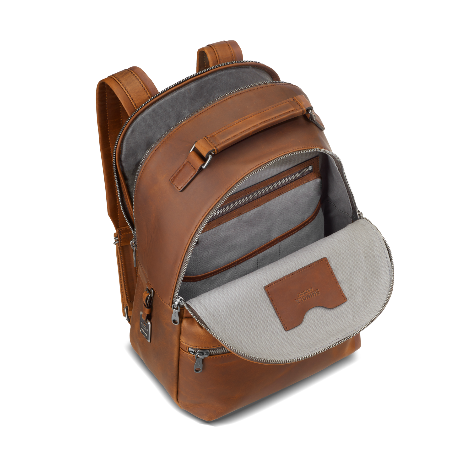 The Shinola Runwell Backpack in medium brown leather is shown open against a white background, featuring a gray fabric interior with multiple compartments, zippered pockets, and an interior leather patch.