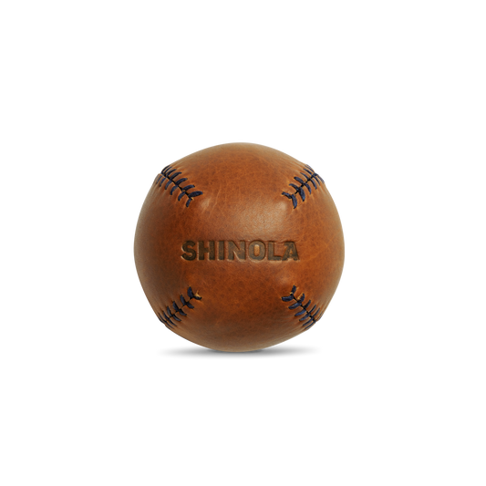 A brown premium leather baseball with visible stitching and "Shinola" embossed, the Leather Baseball - Bourbon / Navy exemplifies Shinola’s expert craftsmanship, set against a clean white background.