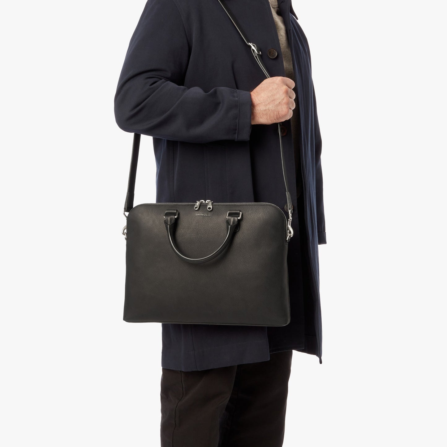 A person in a dark coat holds the Shinola Portfolio Briefcase - Black with a long strap near their chest against a plain white background.