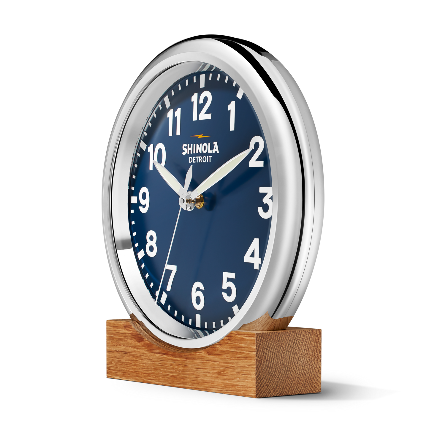 The 14" Runwell Clock - Navy by Shinola features a round silver frame, blue face, white numbers and hands. "Shinola Detroit" is printed on the dial. The clock rests on a wooden base and is set against a plain white background.
