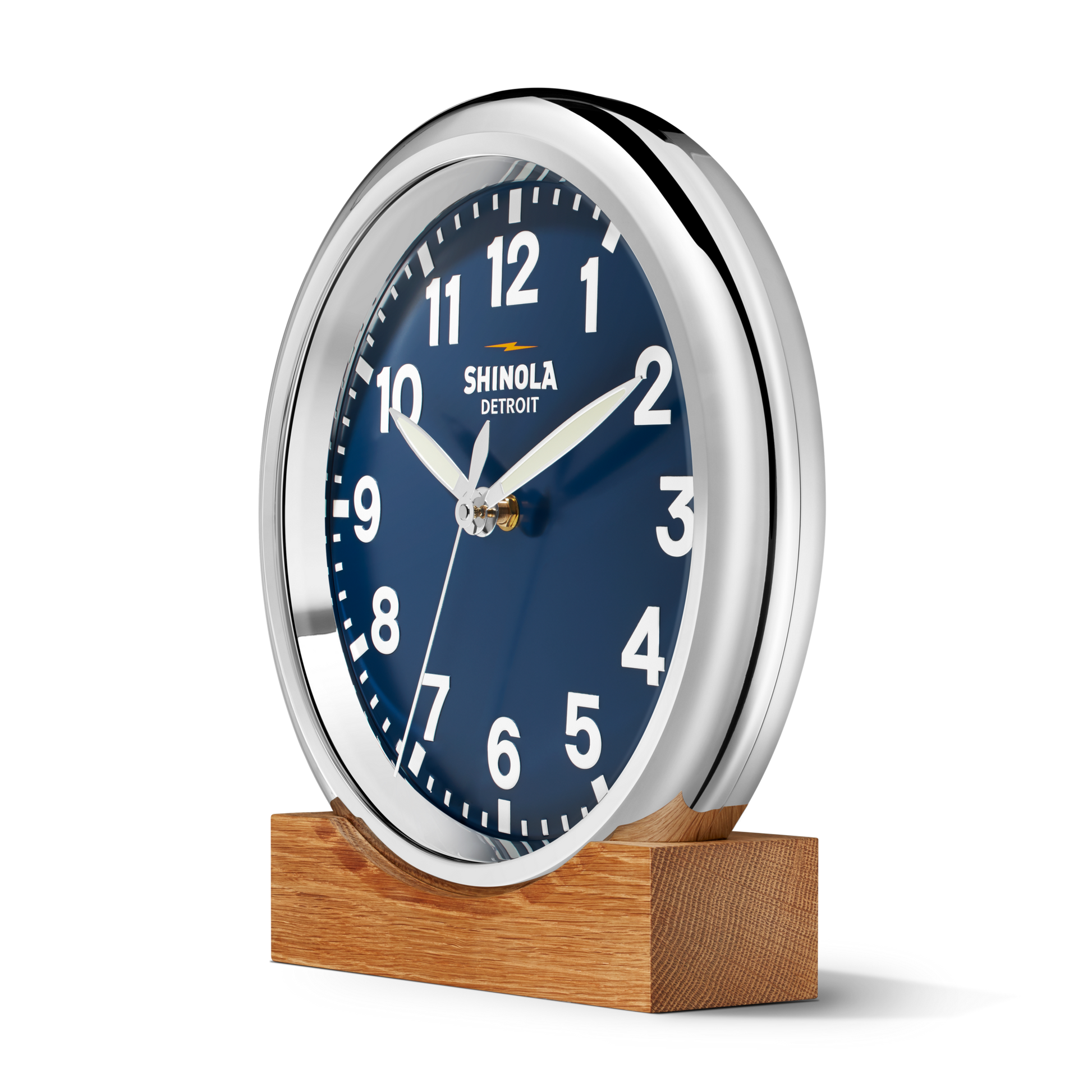 The 14" Runwell Clock - Navy by Shinola features a round silver frame, blue face, white numbers and hands. "Shinola Detroit" is printed on the dial. The clock rests on a wooden base and is set against a plain white background.