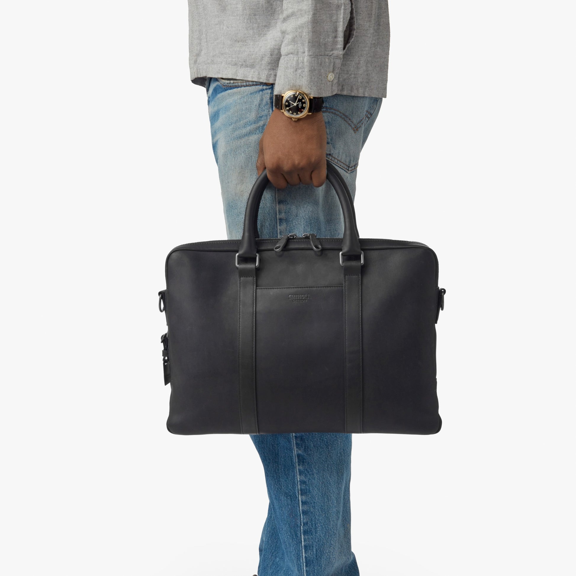 A person in a gray shirt, blue jeans, and a watch holds the shinola-jp.computer Briefcase - Black by its handles against a plain white background.