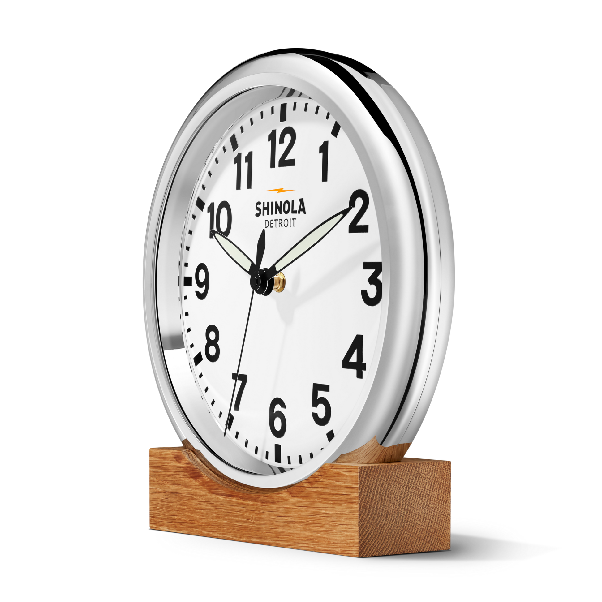 A round, silver-framed 14" Runwell Clock - White by Shinola with black numbers and hands shows 10:09 on a wooden stand. The clock face reads Shinola against a plain white background.