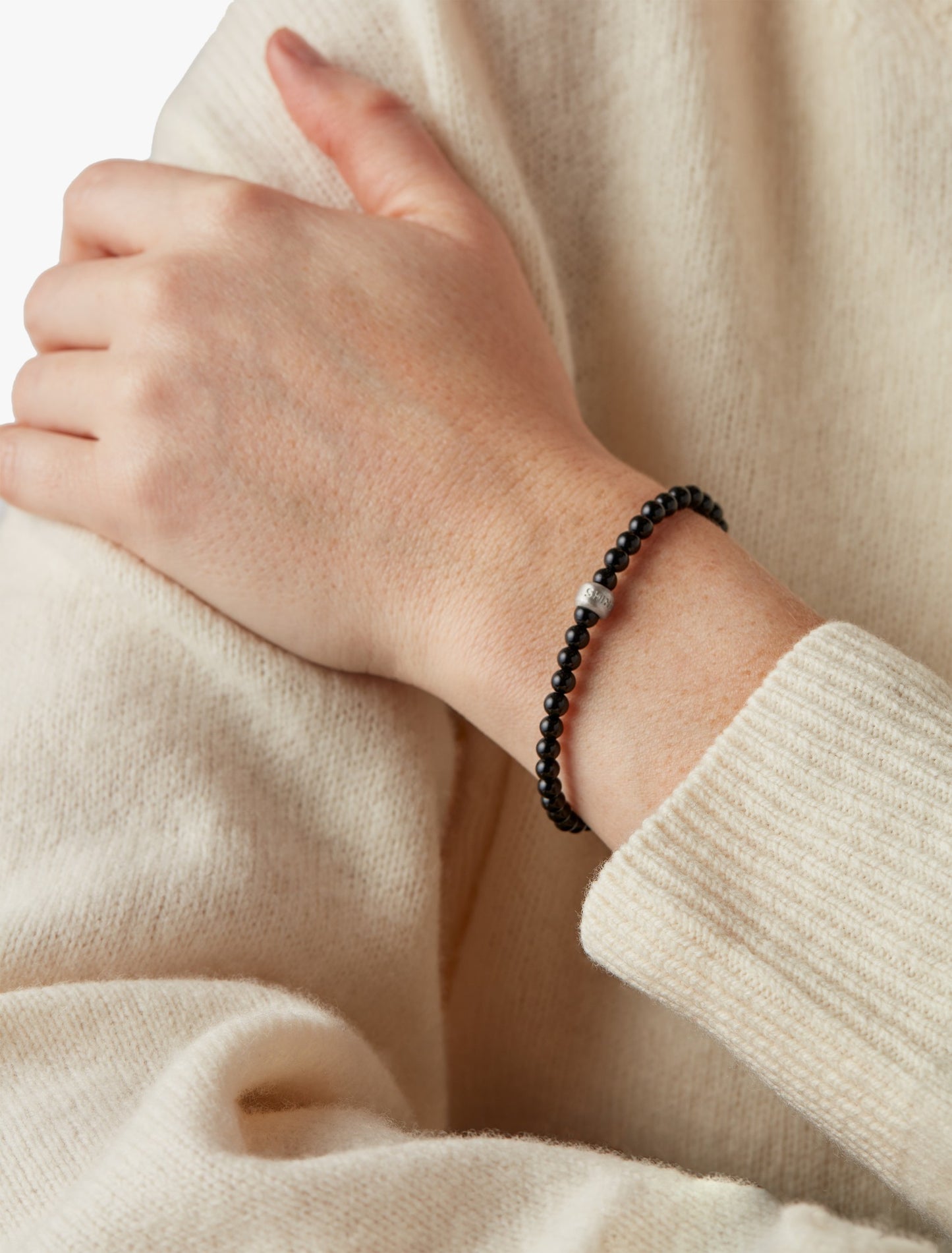 A person in a cream sweater showcases their wrist adorned with the Shinola 4mm Beaded Onyx Bracelet, featuring onyx beads and a sterling silver accent.