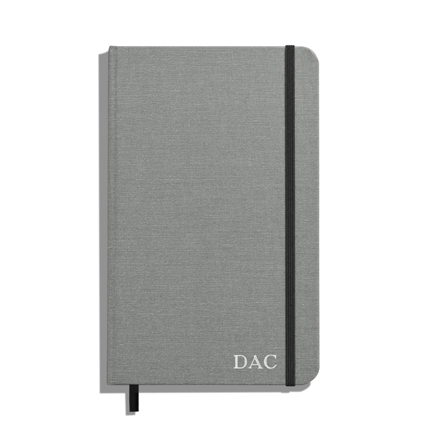 The Shinola Medium Hard Ruled Journal - Stone features a gray hardcover, smyth-sewn binding, black elastic band and ribbon bookmark, with DAC initials in white on the lower right corner of the cover.